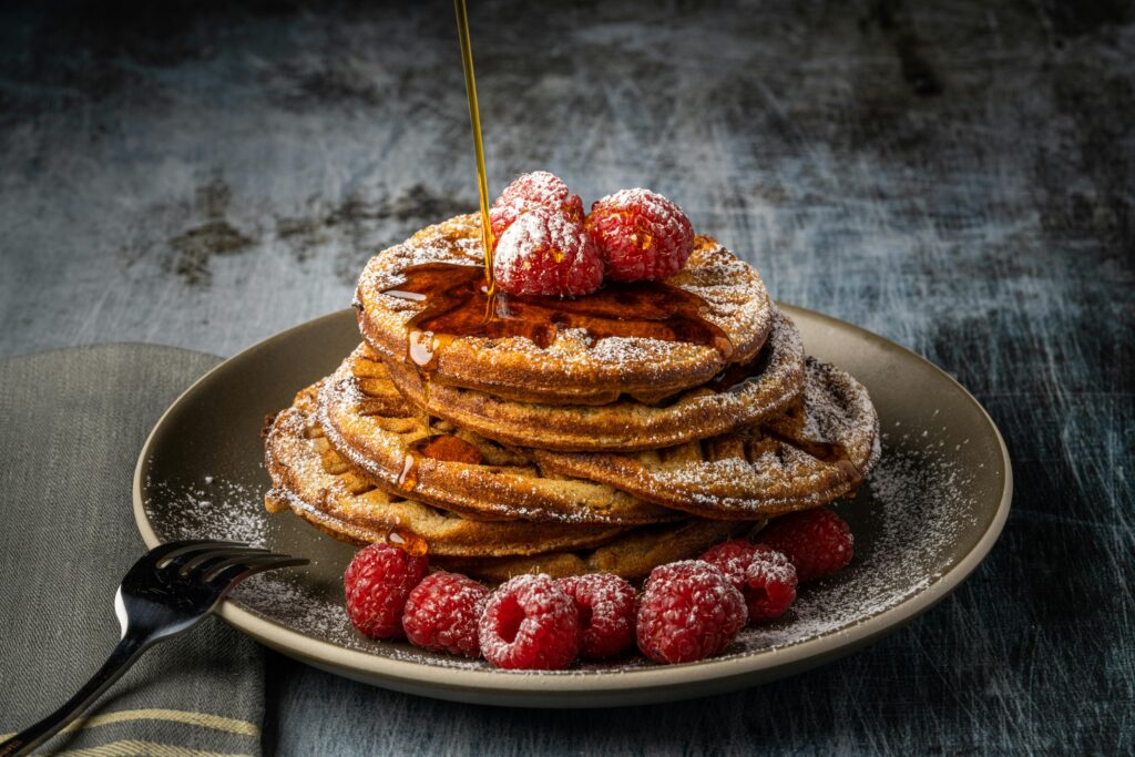 Stack of pancakes topped with raspberries and drizzled with syrup, dusted with icing sugar.